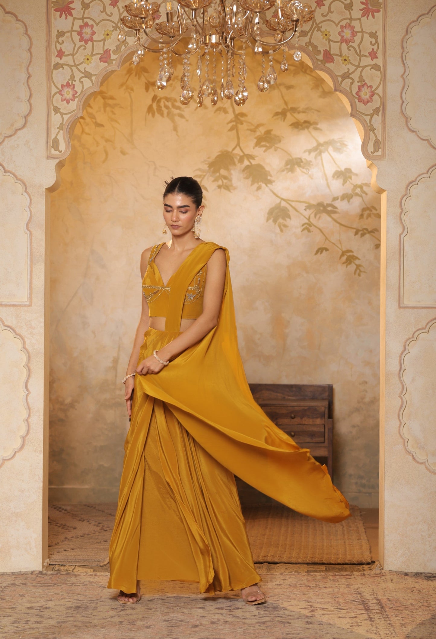 Woman in a mustard yellow saree standing in an ornate room with a chandelier. Golden yellow crepe saree, haldi ceremony saree, embroidered festive saree, designer ethnic wear, elegant haldi outfit, Sunanta Madaan