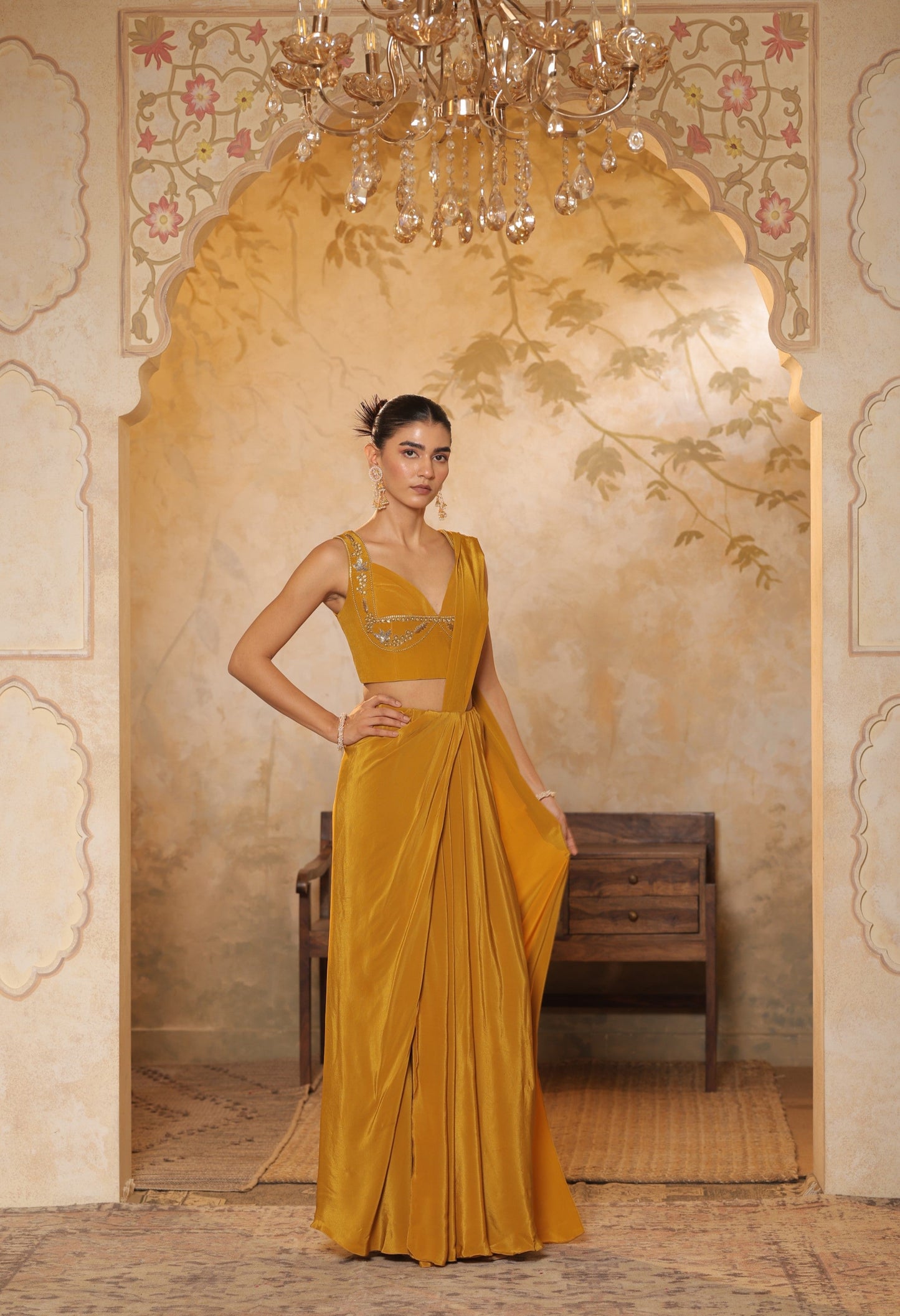 Woman in a mustard yellow saree standing in an ornate room with a chandelier. Golden yellow crepe saree, haldi ceremony saree, embroidered festive saree, designer ethnic wear, elegant haldi outfit, Sunanta Madaan