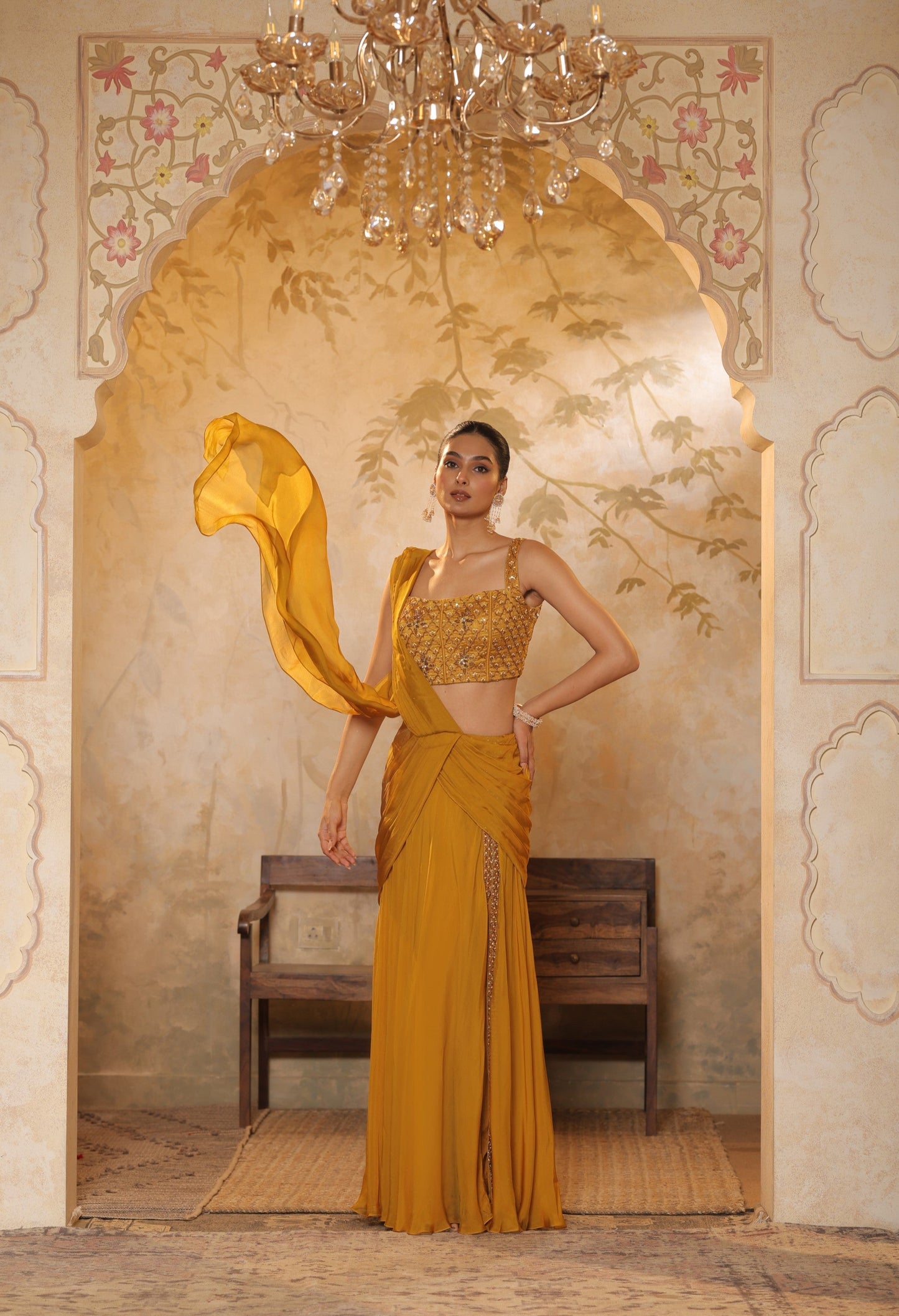 Woman in a mustard yellow traditional outfit standing in an ornate room with a chandelier. Golden yellow saree, chiffon embroidered saree, elegant traditional saree, festive ethnic wear, designer saree online, Sunanta Madaan