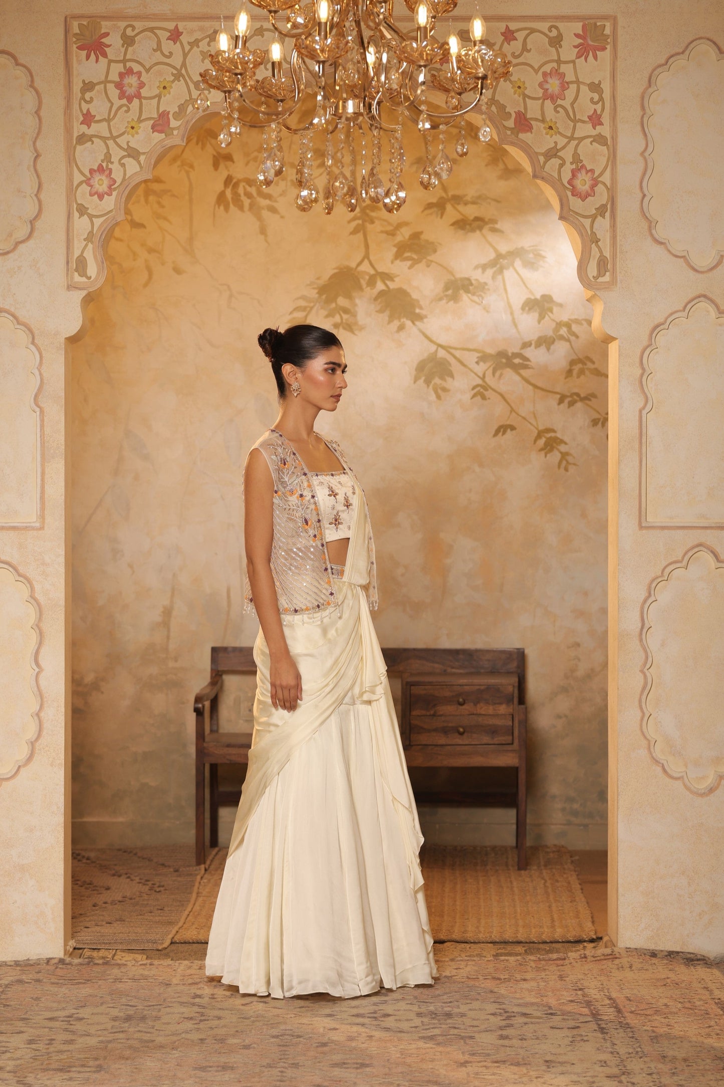 Woman in a traditional outfit standing in an ornate room with a chandelier. ivory georgette saree, saree with embroidered cape, designer wedding saree India, Indo Western bridal outfit, elegant ivory saree, Sunanta Madaan