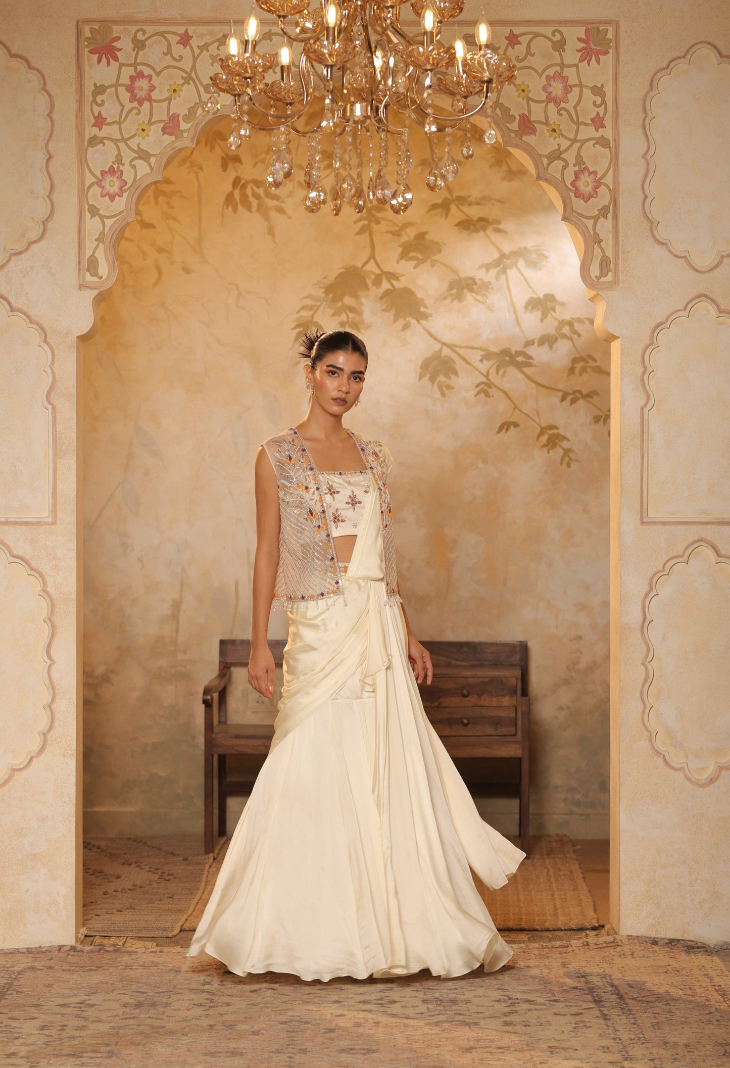 Woman in a traditional outfit standing in an ornate room with a chandelier. ivory georgette saree, saree with embroidered cape, designer wedding saree India, Indo Western bridal outfit, elegant ivory saree, Sunanta Madaan