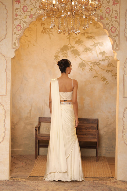 Woman in a white saree standing in a decorated room with a chandelier. ivory georgette saree, saree with embroidered cape, designer wedding saree India, Indo Western bridal outfit, elegant ivory saree, Sunanta Madaan