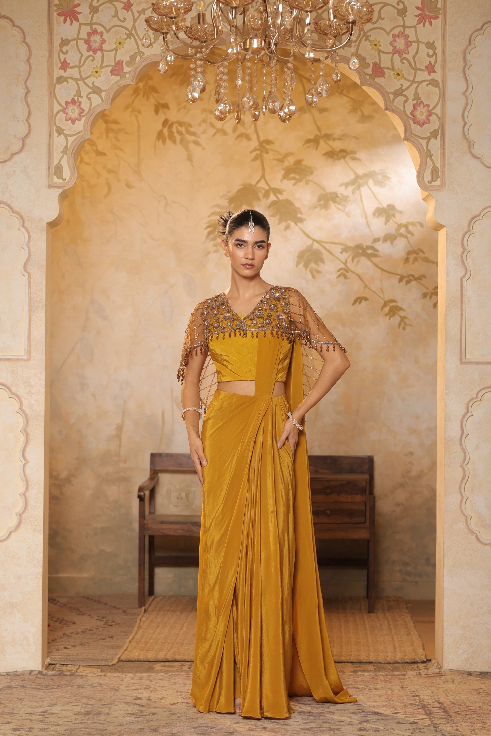 Woman in a mustard yellow saree with intricate blouse design standing in an ornate room. Golden yellow crepe saree, designer cape saree, premium haldi outfit, elegant festive wear, embroidered ethnic saree, Sunanta Madaan