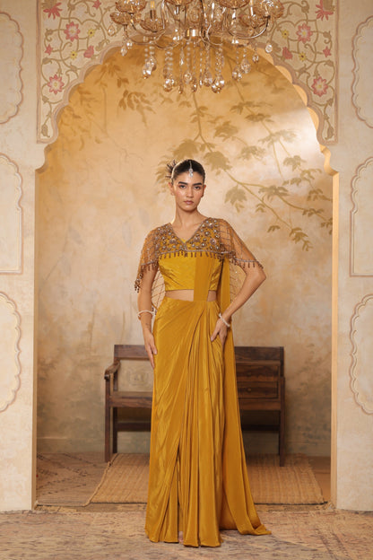 Woman in a mustard yellow saree with intricate blouse design standing in an ornate room. Golden yellow crepe saree, designer cape saree, premium haldi outfit, elegant festive wear, embroidered ethnic saree, Sunanta Madaan