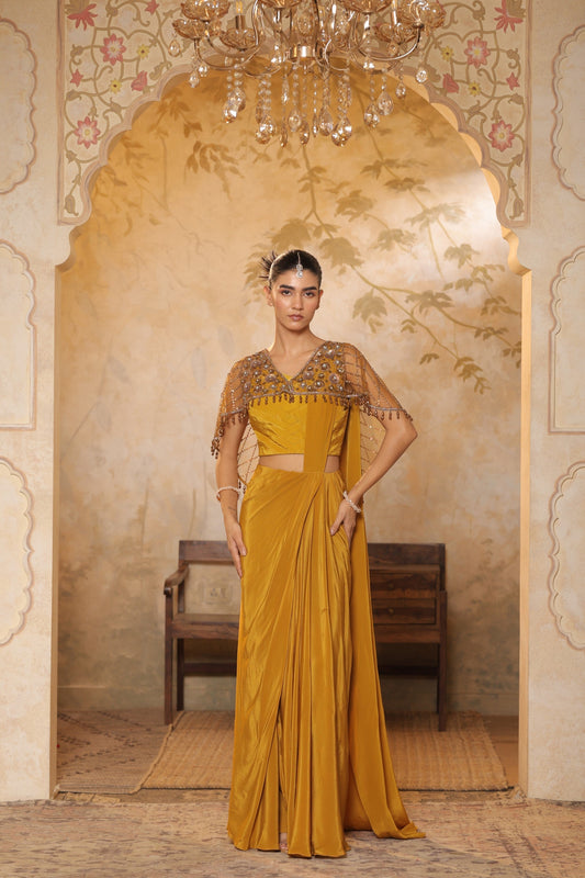 Woman in a mustard yellow saree with intricate blouse design standing in an ornate room. Golden yellow crepe saree, designer cape saree, premium haldi outfit, elegant festive wear, embroidered ethnic saree, Sunanta Madaan