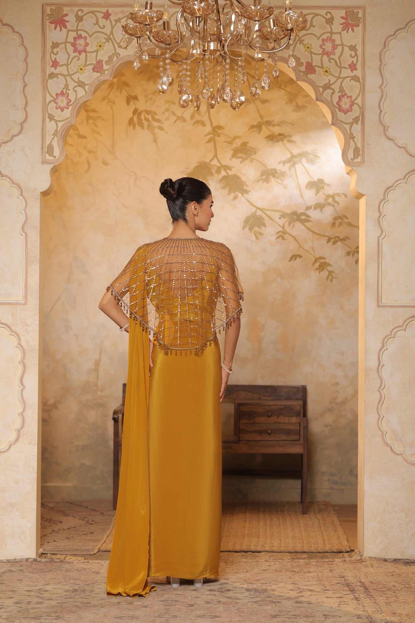 Woman in a mustard yellow dress with intricate detailing standing in a decorated room. Golden yellow crepe saree, designer cape saree, premium haldi outfit, elegant festive wear, embroidered ethnic saree, Sunanta Madaan