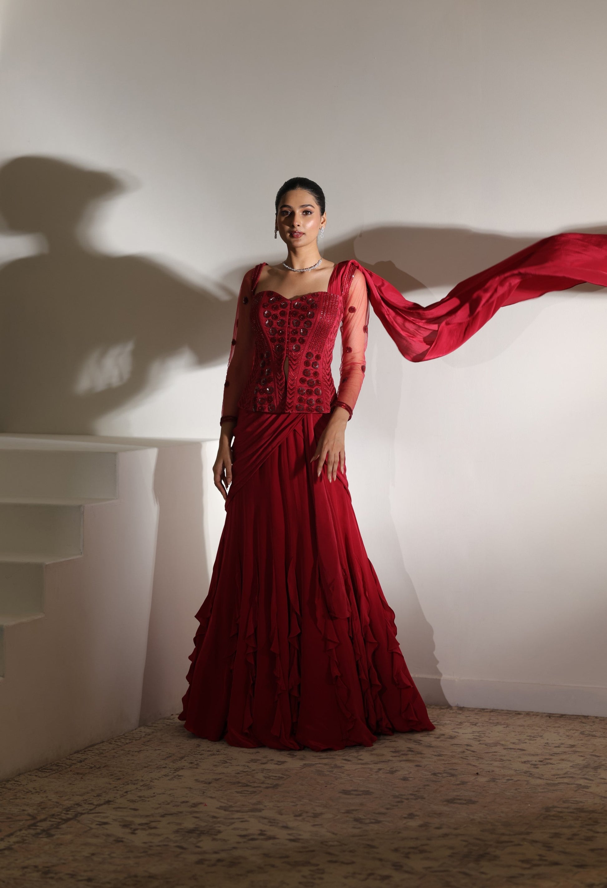 Woman in a red saree with a long train against a white wall; Sunanta Madaan – Burgundy Raw Silk & Crepe Embroidered Corset Saree, Premium Designer Wear, Elegant Festive Outfit, Boho Indo-Western Dress, Indo-Western Fusion Wear, Indo-Western Attire for Engagement, Indo-Western Outfit for Sangeet, Indo-Western Draped & Embroidered Saree, Women’s Designer Saree, Mumbai, Delhi, Hyderabad, Bangalore, Pune, near mev