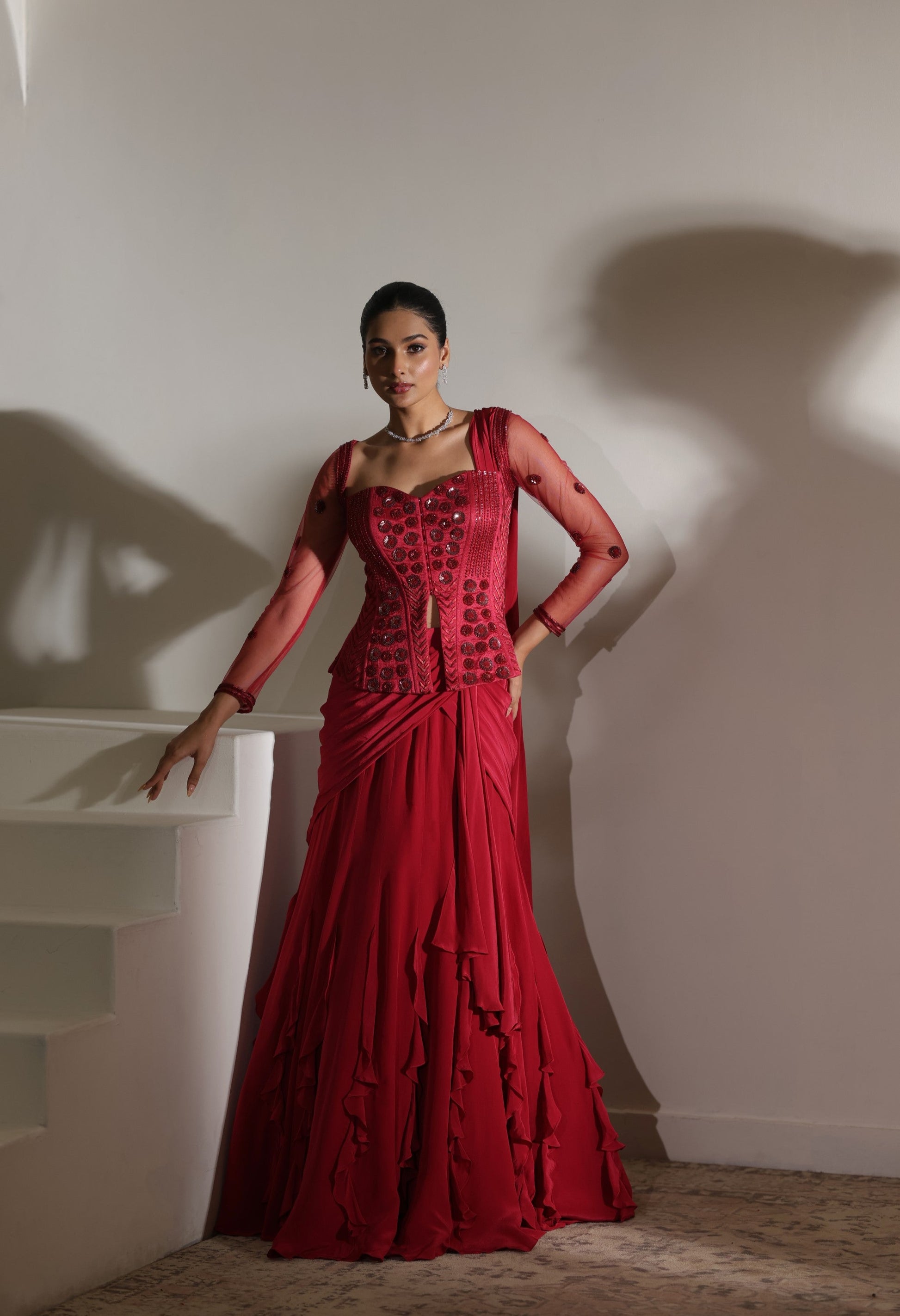 Woman in a red dress standing against a white wall; Sunanta Madaan – Burgundy Raw Silk & Crepe Embroidered Corset Saree, Premium Designer Wear, Elegant Festive Outfit, Boho Indo-Western Dress, Indo-Western Fusion Wear, Indo-Western Attire for Engagement, Indo-Western Outfit for Sangeet, Indo-Western Draped & Embroidered Saree, Women’s Designer Saree, Mumbai, Delhi, Hyderabad, Bangalore, Pune, near me
