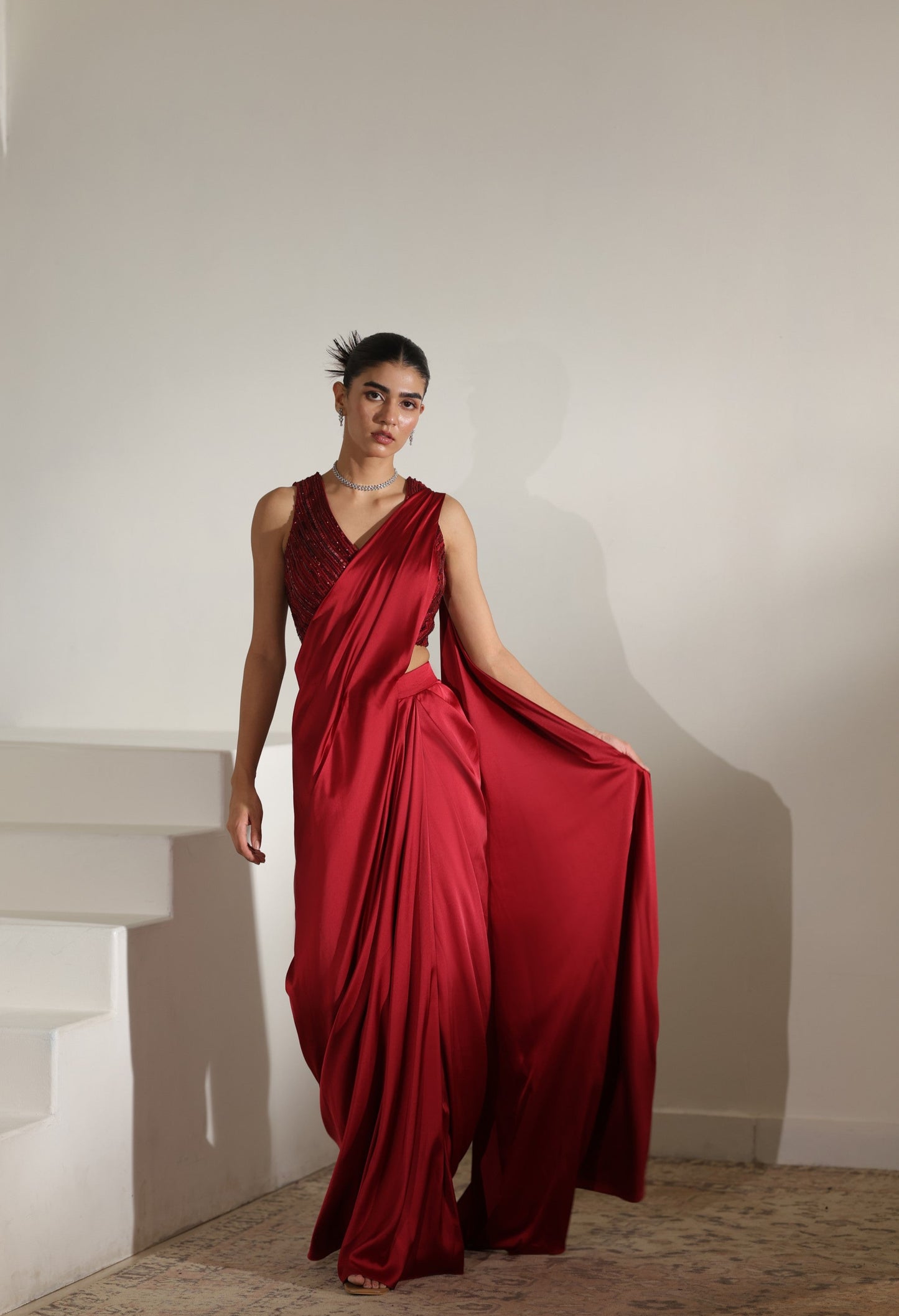 Woman wearing a red draped dress against a plain wall. Sunanta Madaan – Maroon Satin Embroidered Draped Saree, Elegant Wedding Wear, Premium Festive Outfit, Boho Indo-Western Dress, Indo-Western Fusion Wear, Indo-Western Attire for Engagement, Indo-Western Outfit for Sangeet, Indo-Western Draped & Embroidered Saree, Women’s Designer Bridal Ensemble, Delhi, Jaipur, Hyderabad, Ahmedabad, Bangalore, near me