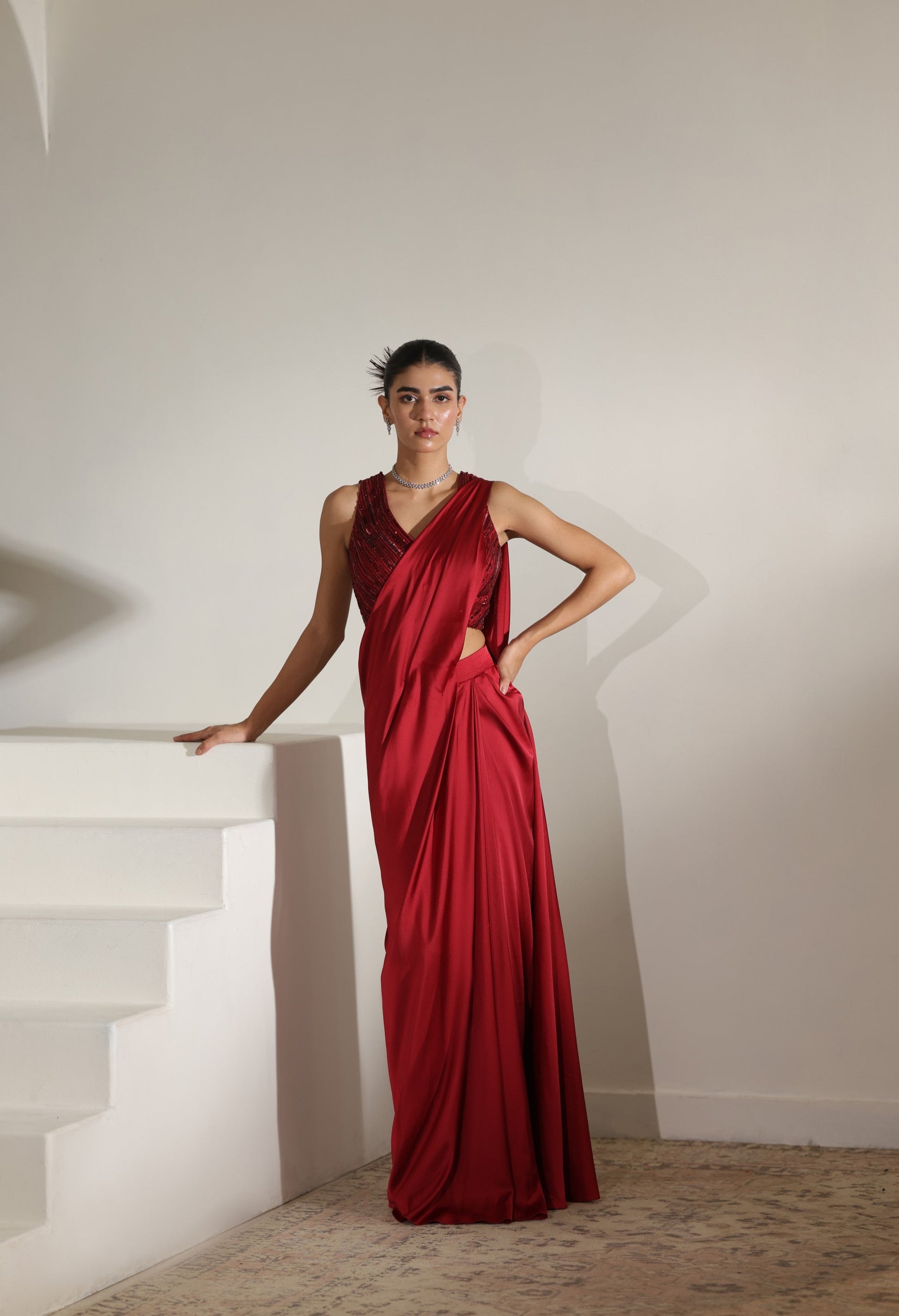 Woman in a red evening gown standing on a staircase with a plain wall background; Sunanta Madaan – Maroon Satin Embroidered Draped Saree, Elegant Wedding Wear, Premium Festive Outfit, Boho Indo-Western Dress, Indo-Western Fusion Wear, Indo-Western Attire for Engagement, Indo-Western Outfit for Sangeet, Indo-Western Draped & Embroidered Saree, Women’s Designer Bridal Ensemble, Delhi, Jaipur, Hyderabad, Ahmedabad, Bangalore, near me