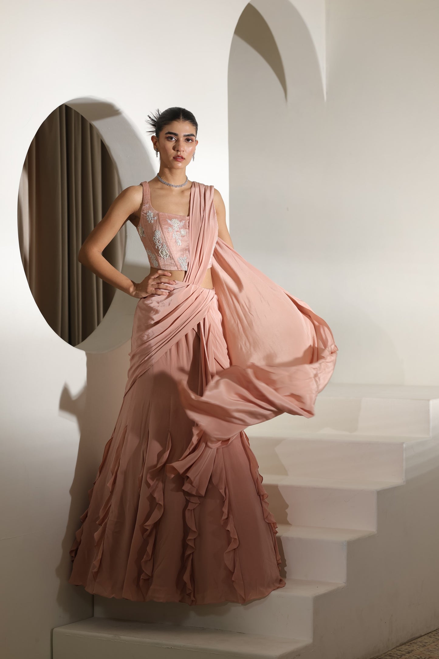 Woman in a pink evening gown standing in a softly lit room with architectural elements. Sunanta Madaan – Mocha Mousse Crepe Embroidered Drape Lehenga Saree, Premium Festive Wear, Elegant Party Outfit, Boho Indo-Western Dress, Indo-Western Fusion Wear, Indo-Western Attire for Engagement, Indo-Western Outfit for Sangeet, Indo-Western Draped & Embroidered Skirt, Women’s Designer Festive Ensemble, Delhi, Jaipur, Hyderabad, Ahmedabad, Bangalore, near me