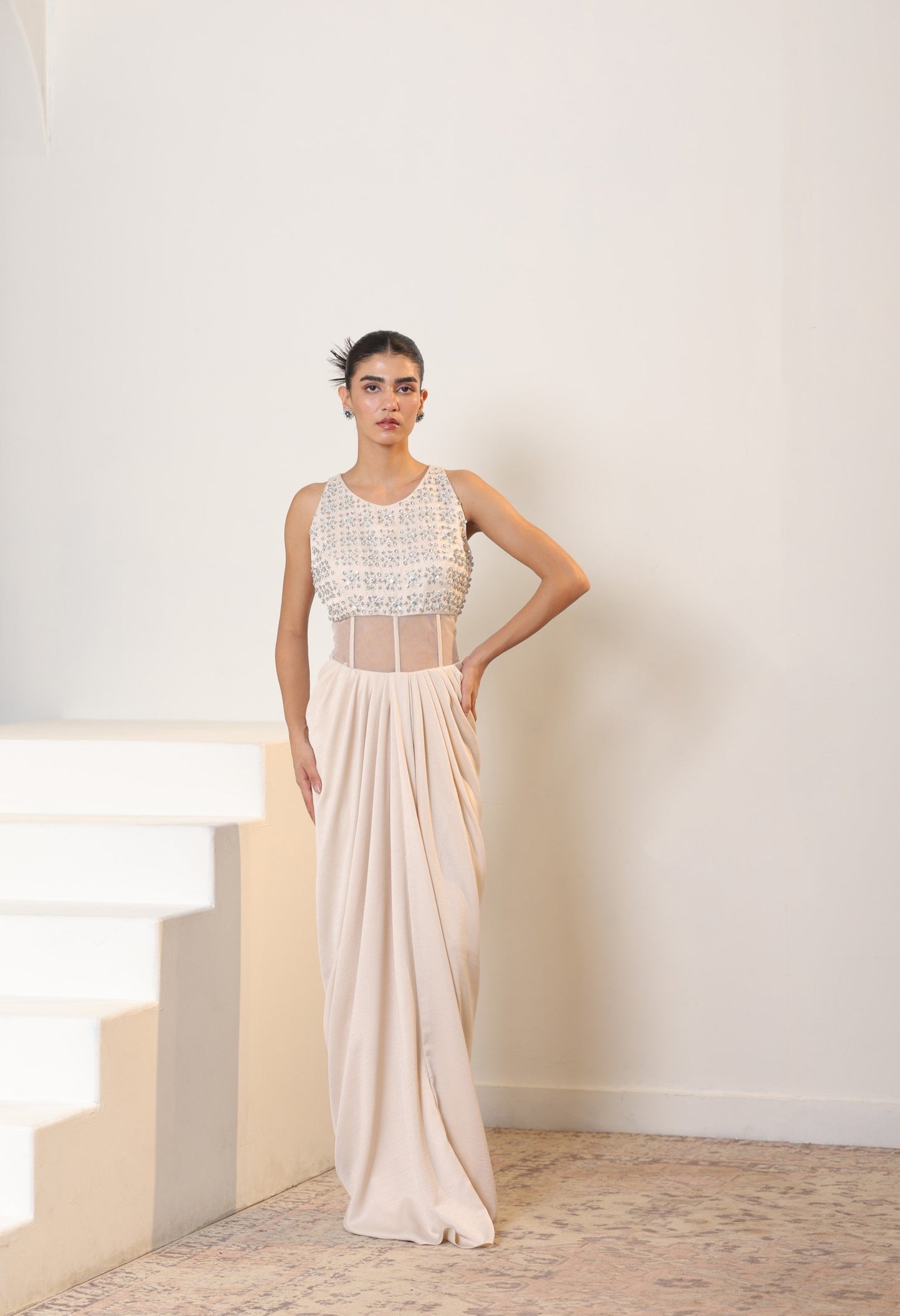 Woman wearing a white dress with intricate detailing in a minimal indoor setting; Sunanta Madaan – Ivory Crepe Satin Western Embroidered Gown, Premium Cocktail Wear, Elegant Party Outfit, Boho Indo-Western Dress, Indo-Western Fusion Wear, Women’s Formal Evening Dress, Indo-Western Outfit for Reception, Party Outfit, Indo-Western Draped Dress, Jaipur, Delhi, Hyderabad, Ahmedabad, Bangalore, near me