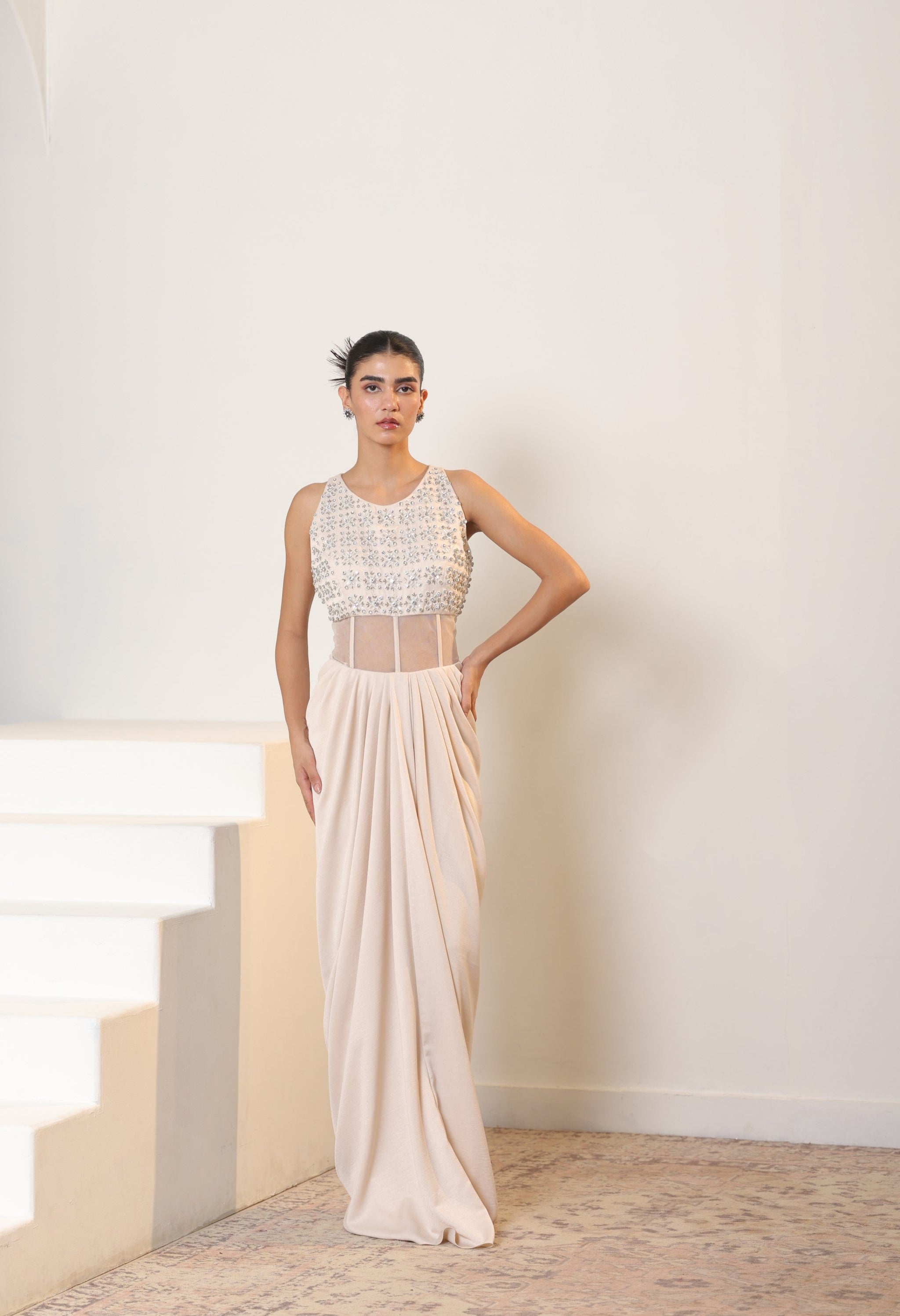 Woman wearing a white dress with intricate detailing in a minimal indoor setting; Sunanta Madaan – Ivory Crepe Satin Western Embroidered Gown, Premium Cocktail Wear, Elegant Party Outfit, Boho Indo-Western Dress, Indo-Western Fusion Wear, Women’s Formal Evening Dress, Indo-Western Outfit for Reception, Party Outfit, Indo-Western Draped Dress, Jaipur, Delhi, Hyderabad, Ahmedabad, Bangalore, near me