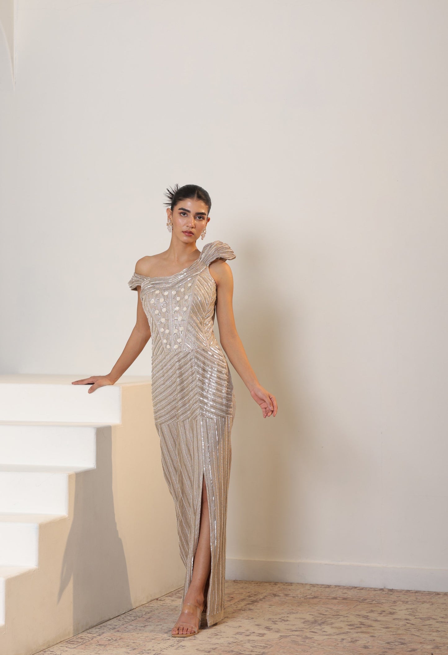 Woman in a silver one-shoulder evening gown standing on a staircase. Sunanta Madaan – Champagne Metallic Lycra Crepe Embroidered Gown, Elegant Cocktail Wear, Evening Party Dress, Boho Indo-Western Dress, Indo-Western Fusion Wear, Women’s Formal Gown, Indo-Western Outfit for Reception, Party Outfit, Indo-Western Draped Dress, Jaipur, Delhi, Pune, Ahmedabad, Hyderabad, near me