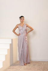 Woman in a silver one-shoulder gown standing in a minimalistic room. Sunantamadaan Saree, pre draped saree, peach saree, simple saree, latest saree design, sarees online, pink saree, satin saree, ready made saree, white colour saree, fancy saree, party wear saree, Sunanta Madaan – Pink Metallic Lycra Crepe Saree, Premium Reception Wear, Elegant Party Outfit, Boho Indo-Western Dress, Indo-Western Fusion Wear, Women’s Formal Evening Dress, Indo-Western Outfit for Reception, Party Outfit, Indo-Western Draped &
