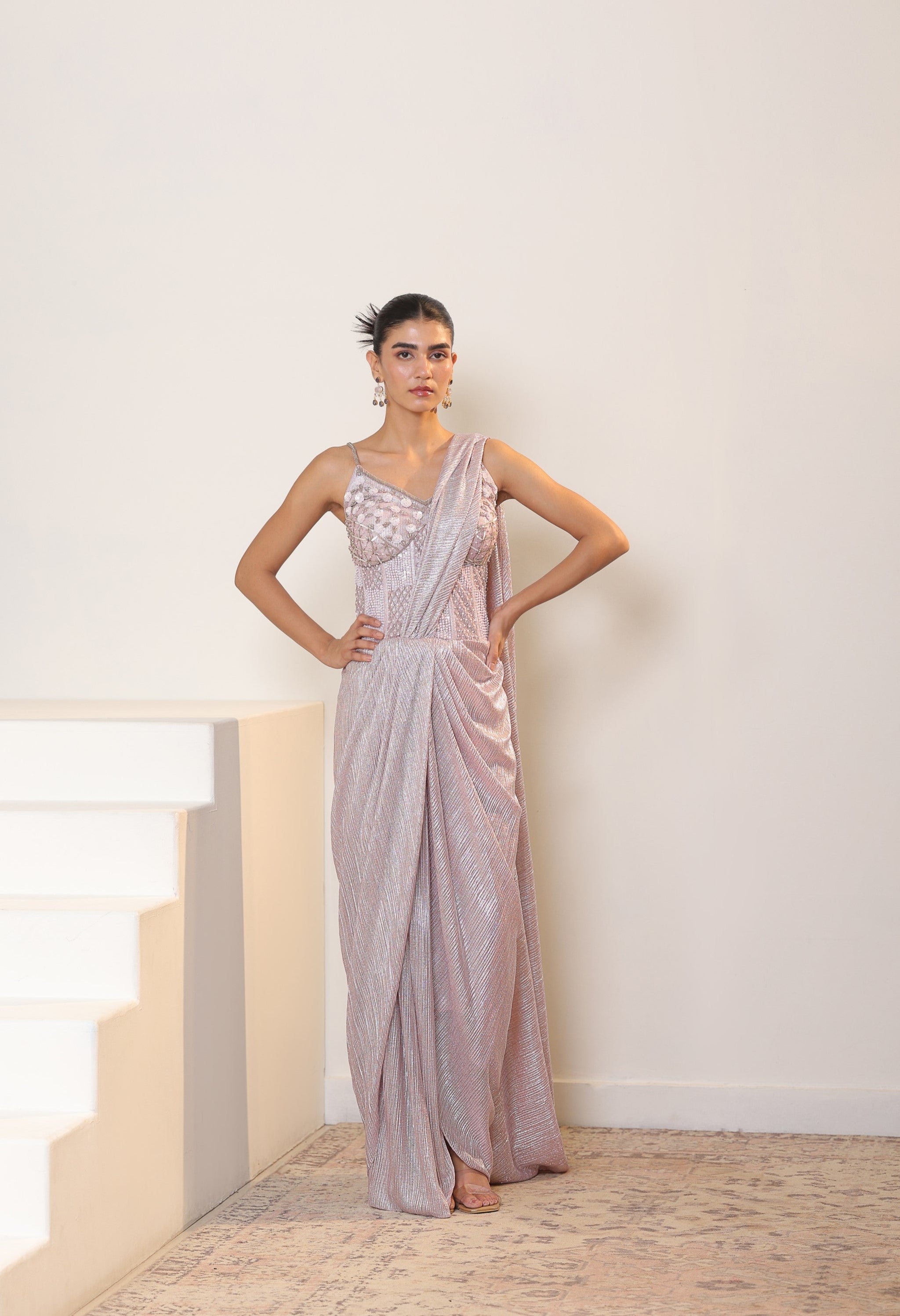 Woman in a silver one-shoulder gown standing in a minimalistic room. Sunantamadaan Saree, pre draped saree, peach saree, simple saree, latest saree design, sarees online, pink saree, satin saree, ready made saree, white colour saree, fancy saree, party wear saree, Sunanta Madaan – Pink Metallic Lycra Crepe Saree, Premium Reception Wear, Elegant Party Outfit, Boho Indo-Western Dress, Indo-Western Fusion Wear, Women’s Formal Evening Dress, Indo-Western Outfit for Reception, Party Outfit, Indo-Western Draped &