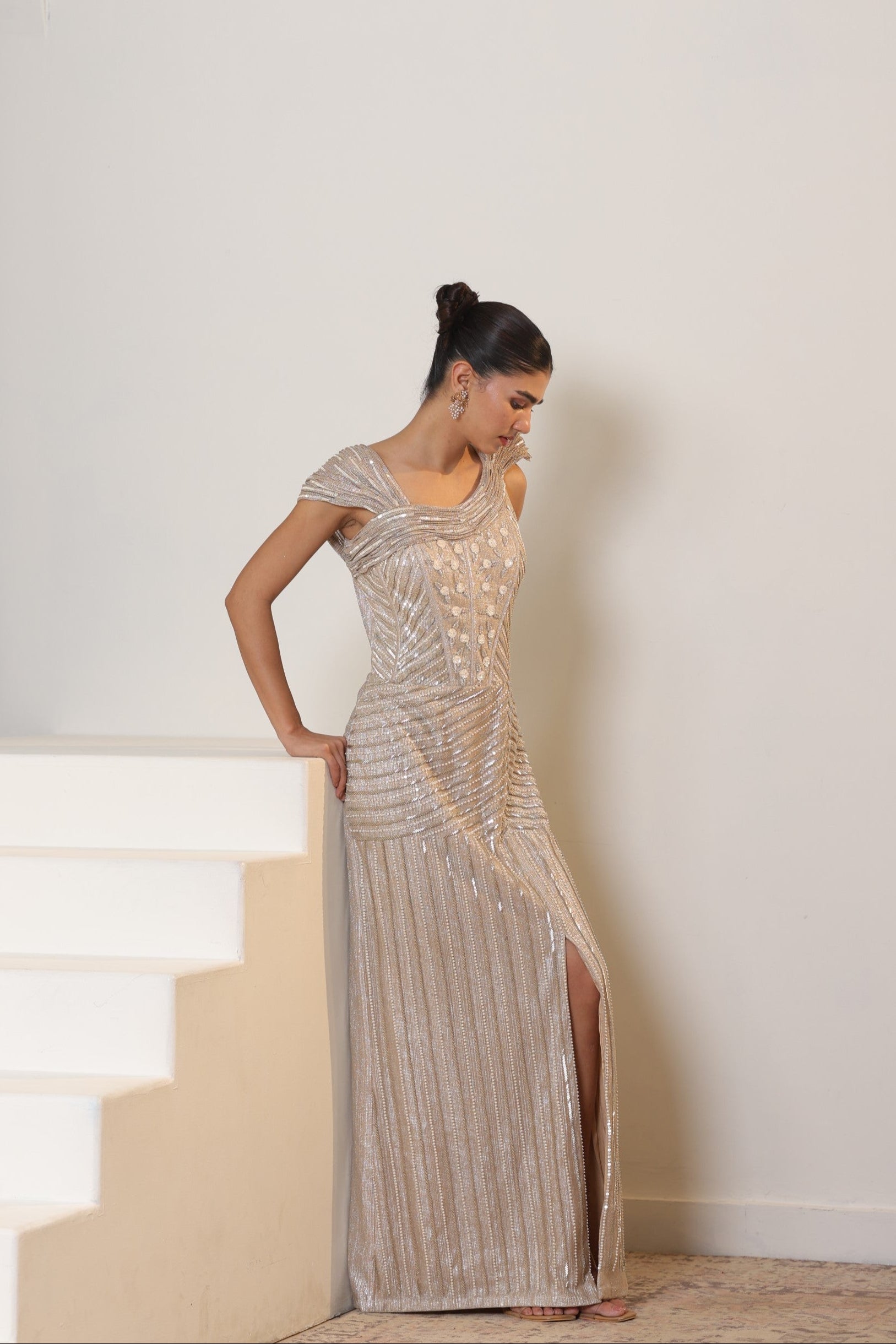 Woman in a long, elegant dress standing on a staircase with a plain wall background; Sunanta Madaan – Champagne Metallic Lycra Crepe Embroidered Gown, Elegant Cocktail Wear, Evening Party Dress, Boho Indo-Western Dress, Indo-Western Fusion Wear, Women’s Formal Gown, Indo-Western Outfit for Reception, Party Outfit, Indo-Western Draped Dress, Jaipur, Delhi, Pune, Ahmedabad, Hyderabad, near me