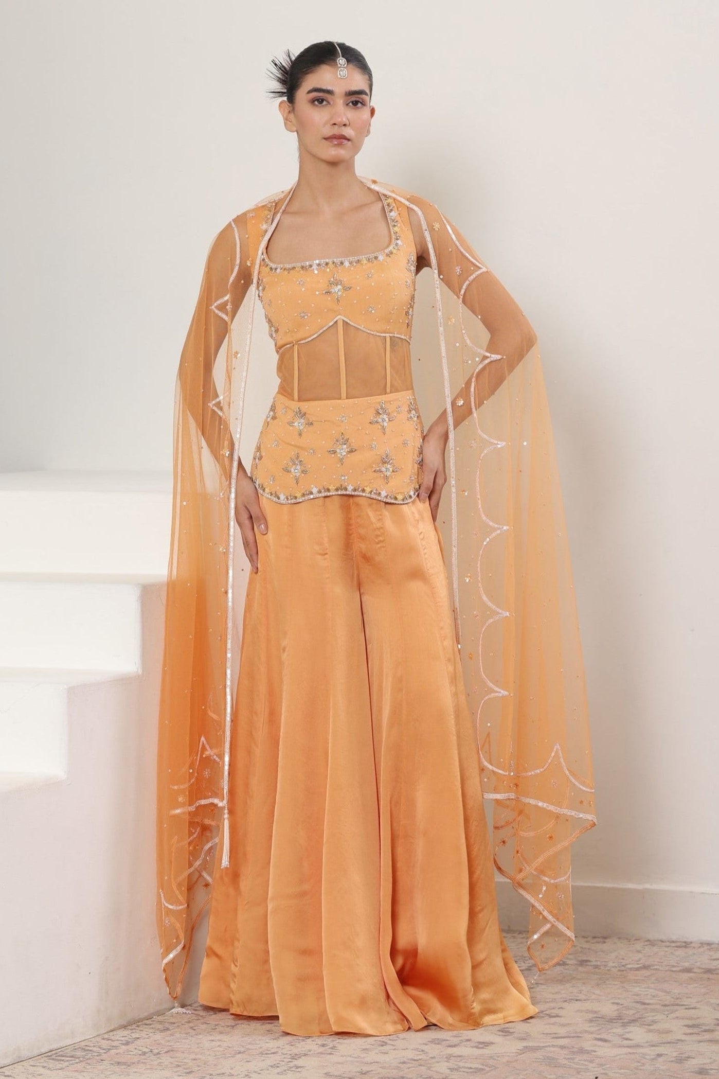 Woman wearing a mustard yellow traditional outfit with a sheer dupatta against a white background; Orange silk crepe sharara, draped festive outfit, elegant ethnic wear, designer traditional attire, premium party ensemble, Sunanta Madaan, near me, Mumbai, Delhi, Hyderabad, Bangalore, Pune