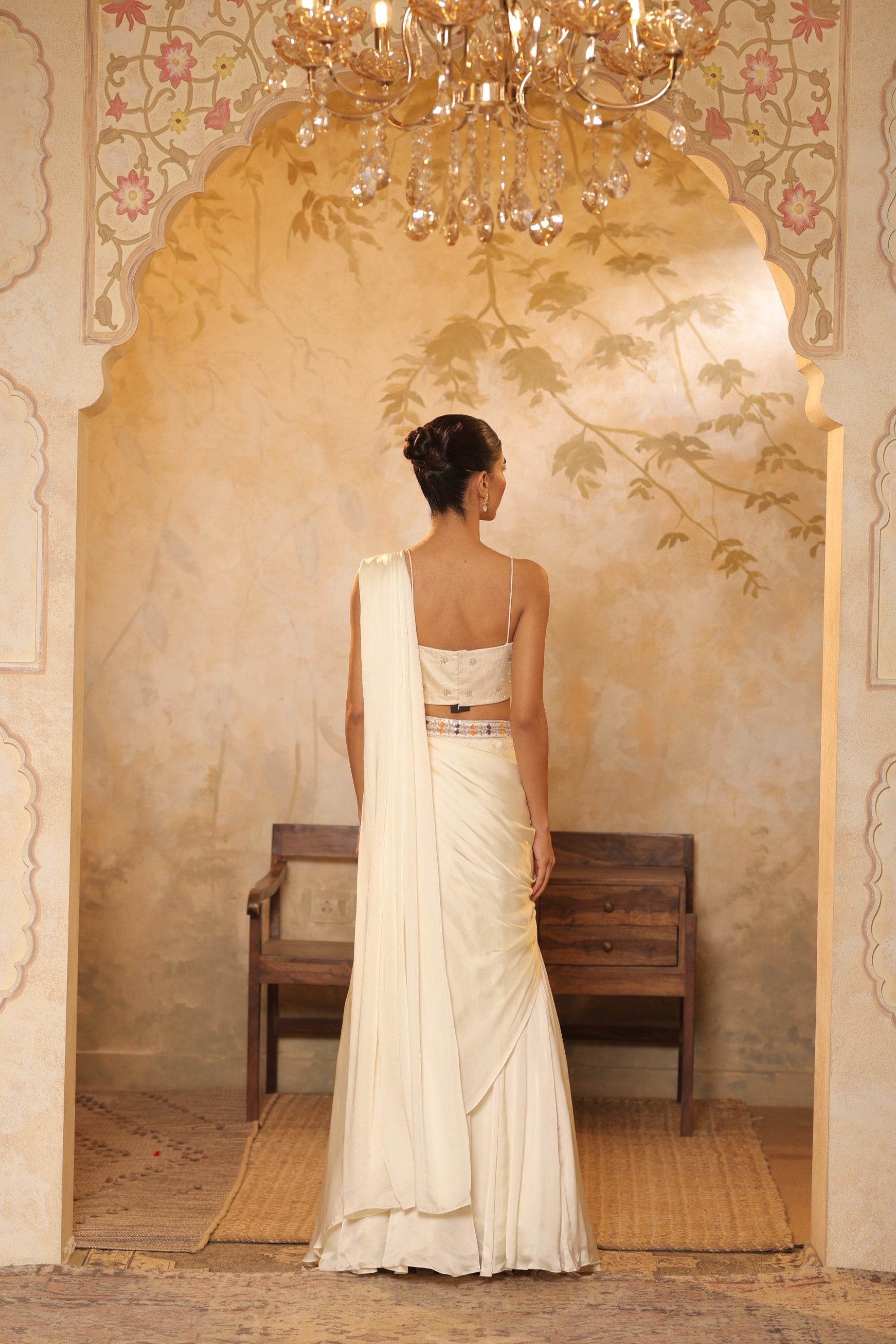 Woman in a white saree standing in a decorated room with a chandelier. ivory georgette saree, saree with embroidered cape, designer wedding saree India, Indo Western bridal outfit, elegant ivory saree, Sunanta Madaan