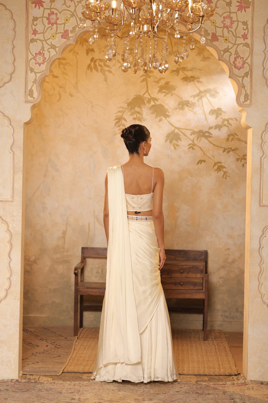 Woman in a white saree standing in a decorated room with a chandelier. ivory georgette saree, saree with embroidered cape, designer wedding saree India, Indo Western bridal outfit, elegant ivory saree, Sunanta Madaan