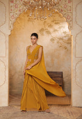 Woman in a mustard yellow saree standing in an ornate room with a chandelier. Golden yellow crepe saree, haldi ceremony saree, embroidered festive saree, designer ethnic wear, elegant haldi outfit, Sunanta Madaan
