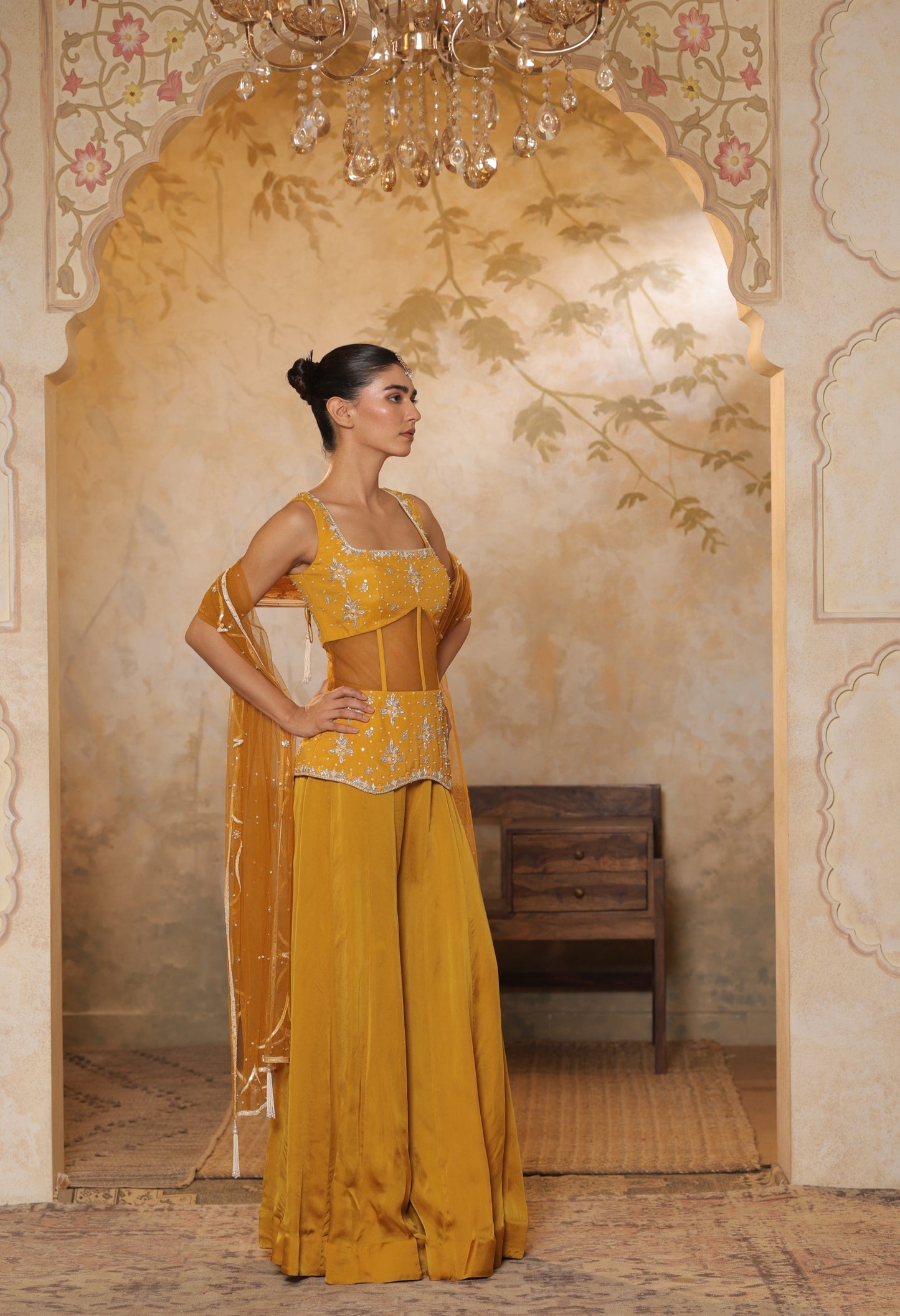 Woman in a mustard yellow traditional outfit standing in an ornate room with a chandelier. Yellow silk crepe sharara, festive ethnic outfit, elegant occasional wear, designer traditional attire, premium party ensemble, Sunanta Madaan, near me, Mumbai, Delhi, Hyderabad, Bangalore, Pune