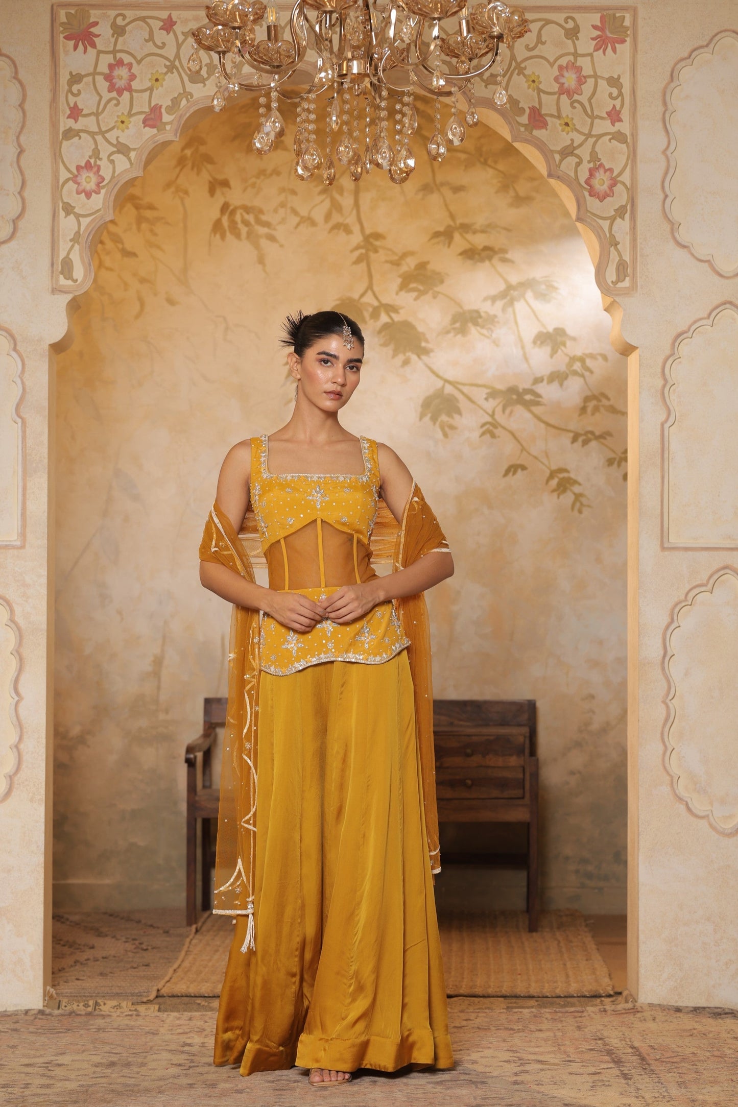 Woman in a yellow traditional outfit standing in an ornate room with a chandelier. Yellow silk crepe sharara, festive ethnic outfit, elegant occasional wear, designer traditional attire, premium party ensemble, Sunanta Madaan, near me, Mumbai, Delhi, Hyderabad, Bangalore, Pune