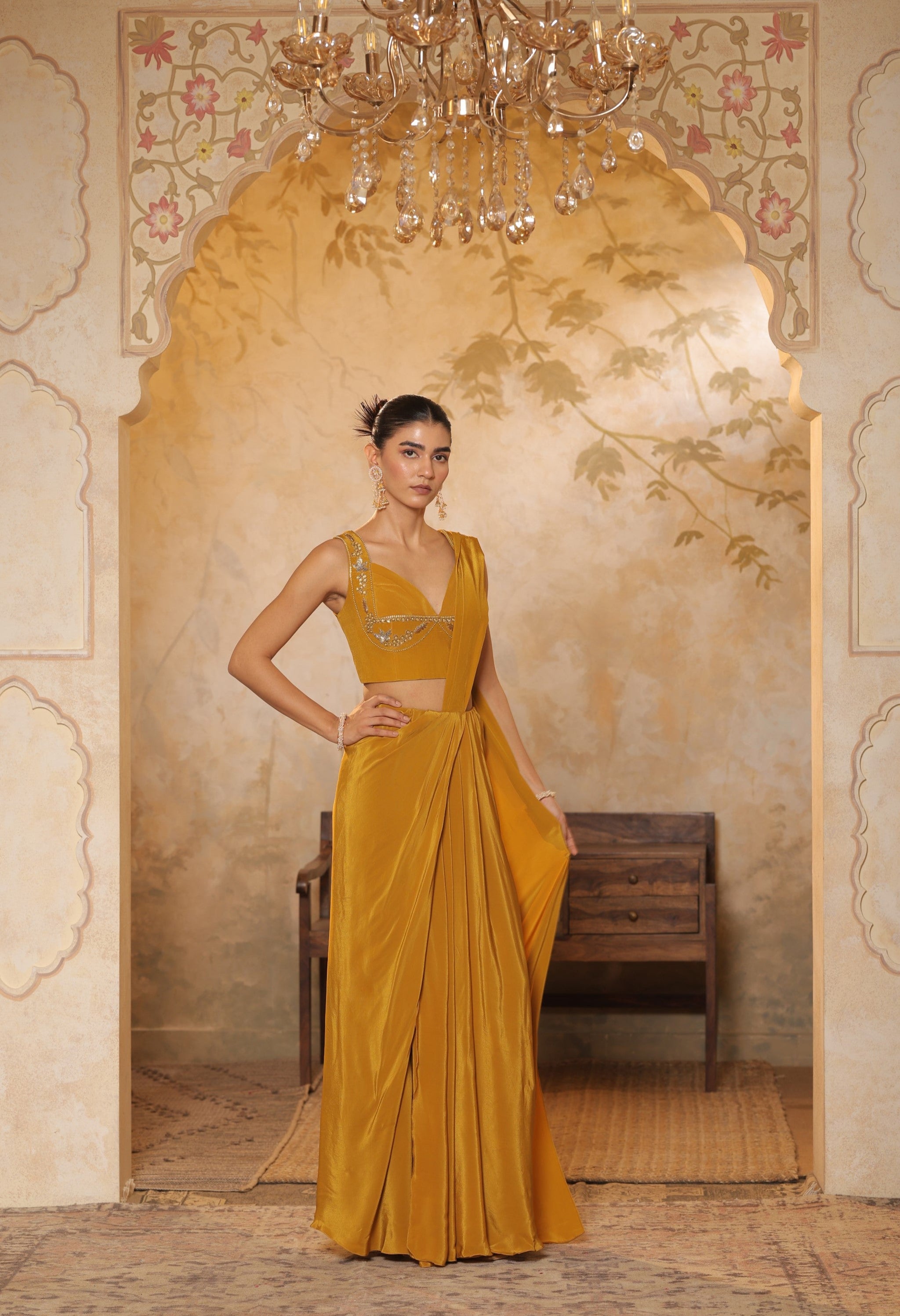 Woman in a mustard yellow saree standing in an ornate room with a chandelier. Golden yellow crepe saree, haldi ceremony saree, embroidered festive saree, designer ethnic wear, elegant haldi outfit, Sunanta Madaan