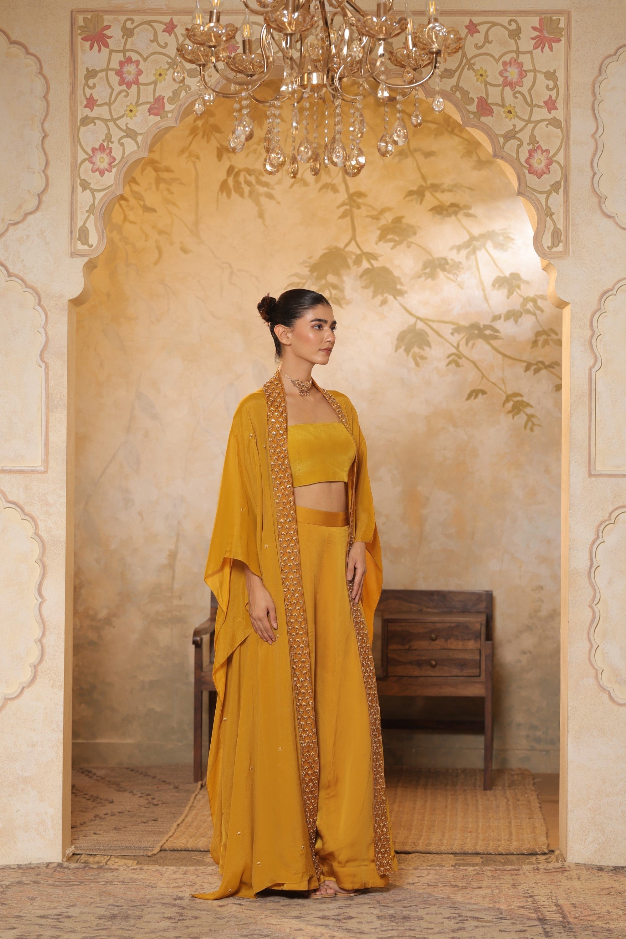 Woman in a yellow traditional outfit standing in an ornate room with a chandelier. Golden yellow sharara set, embroidered wedding outfit, designer festive wear, elegant ethnic ensemble, premium traditional attire, Sunanta Madaan
